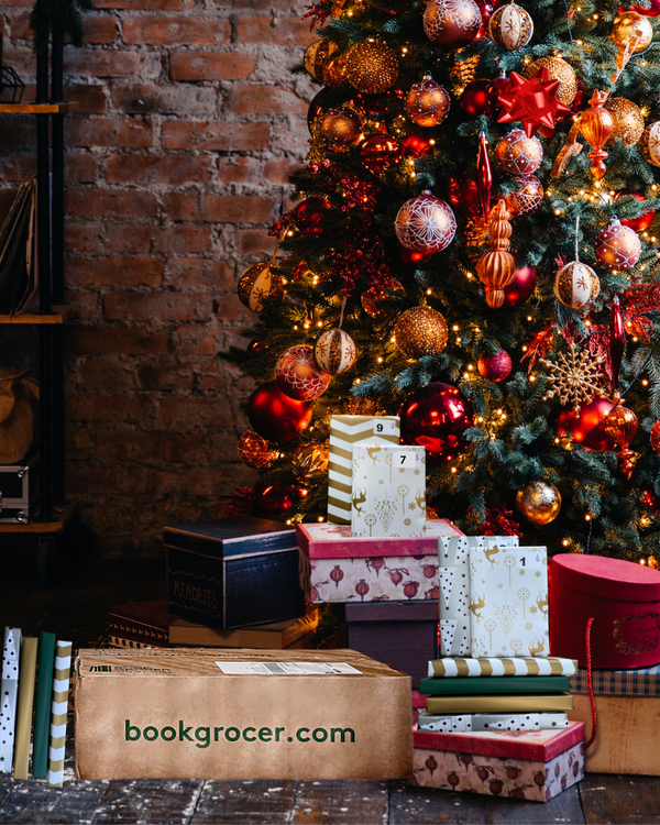 Image shows a Book Grocer Advent Book Box at the bottom of a Christmas tree, surrounded by wrapped books from inside and other Christmas presents.